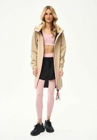 Kobieta w beżowej przeciwdeszczowej kurtce z kapturem, na różowym sportowym biustonoszu i legginsach, czarnych krótkich spodenkach oraz czarnych trampkach, stojąca na białym tle.