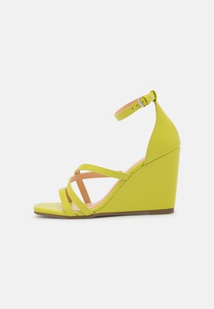 Żółte sandały na koturnie z paskami i klamrą wokół kostki. Wykonane z gładkiego materiału, posiadają teksturowany obcas na koturnie.