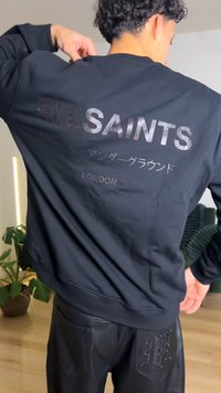 Sudadera negra con los textos "ALL SAINTS" y "LONDON" en la espalda con acabado brillante. La tela tiene una textura suave, típica de una mezcla de algodón.