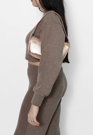 Borsa a spalla in rosè metallizzato con una forma strutturata, chiusura con zip e tracolla regolabile. Indossata con un maglione corto marrone e pantaloni.