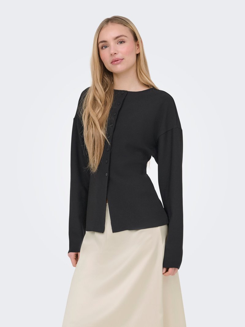 Jeune femme aux cheveux blonds longs portant un pull noir boutonné ajusté et une jupe beige, se tenant devant un fond uni.