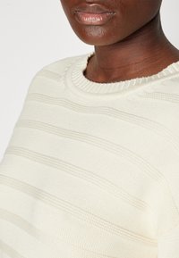 Suéter de punto en color crema claro con rayas texturizadas horizontales. Cuello redondo de canalé y bordes ligeramente deshilachados. Tela suave y lisa.