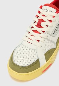 Sneaker med hvit perforert skinn og olivengrønn semsket skinn. Har røde teksturerte lisser, en gul gummisåle og en sølvfarget logo-patch.
