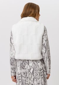 Witte nepbontvest met een kraag, een doorgestikte textuur en een cropped ontwerp, gedragen over een grijs-wit snakenprint outfit.