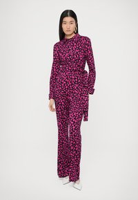 Różowy kombinezon z kwiatowym wzorem, z długimi rękawami, wiązaniem w talii i szerokimi nogawkami, zestawiony z białymi szpilkami i dużymi kolczykami kołami.