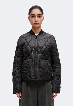 Un mannequin se tient face à l’avant, portant une veste noire matelassée à fermeture éclair et un pantalon foncé, sur un fond clair uni.
