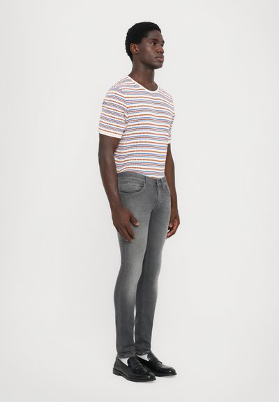 Ριγέ κοντομάνικο πουκάμισο με μπλε και πορτοκαλί γραμμές, συνδυασμένο με στενά γκρι τζιν και μαύρα μοκασίνια, σε ο υδέτερο φόντο.