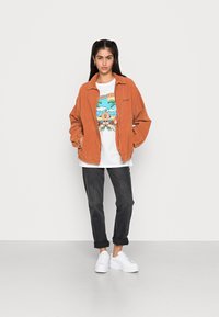 Casaco de corduroy laranja enferrujado sobre uma t-shirt gráfica com design colorido, combinado com jeans escuros e ténis de plataforma brancos.