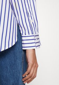 Gros plan sur le bras d'une personne portant une manche de chemise rayée verticale bleue et blanche et un jean bleu sur un fond uni.