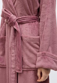 Μαλακή ρόμπα μπάνιου σε απαλό ροζ χρώμα, δεμένη στη μέση με ζώνη, με τσέπες-πατς και μανίκια διπλωμένα στις μανσέτες, που φοράει ένα άτομο.