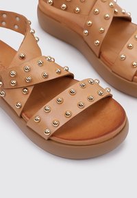 Sandalias de cuero marrón claro con.Clavos de oro, que presentan correas entrelazadas y una suela gruesa y ligera para mayor comodidad. Textura suave y forma redondeada.