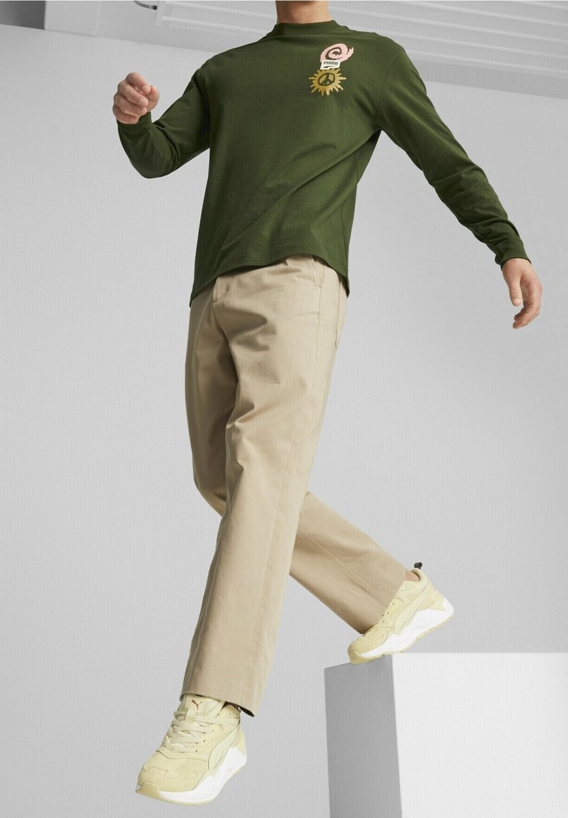 Koszula z długim rękawem w oliwkowym kolorze z nadrukiem graficznym, beżowe spodnie oraz jasne żółte trampki z teksturowanymi akcentami. Model w ruchu na piedestale.