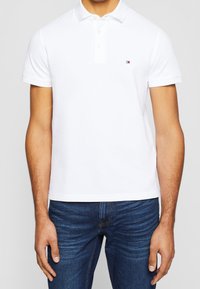 Polo shirt blanc à manches courtes, avec une patte de boutonnage à trois boutons et un petit logo de marque sur la poitrine. Assorti à un jean bleu foncé.