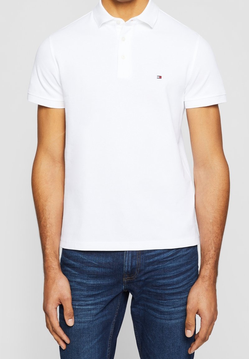 Polo shirt blanc à manches courtes, avec une patte de boutonnage à trois boutons et un petit logo de marque sur la poitrine. Assorti à un jean bleu foncé.