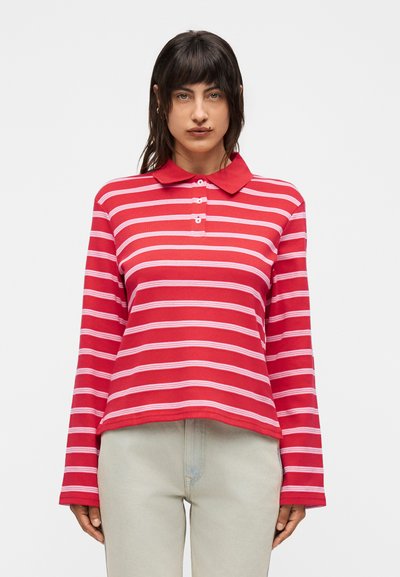 Femme aux cheveux foncés portant un polo à rayures horizontales rouges et blanches à manches longues et un pantalon beige clair, debout devant un fond uni.