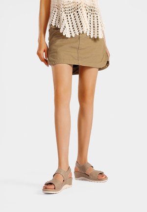 Donna che indossa sandali con zeppa color cuoio, una minigonna verde oliva e un top bianco in crochet, in piedi su sfondo bianco.