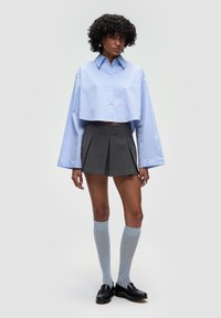 Lyst blå cropped skjorte med knapper og oversized ærmer, parret med en grå plisseret mini-nederdel og knæhøje blå strømper, båret med sorte loafers.