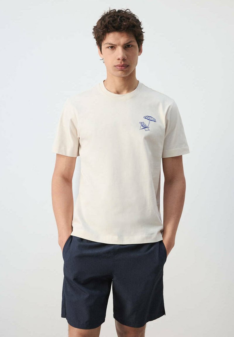 Jeune homme portant un t-shirt beige clair avec un petit dessin de chaise de plage bleue et parasol, associé à un short bleu marine foncé, debout les mains dans les poches.