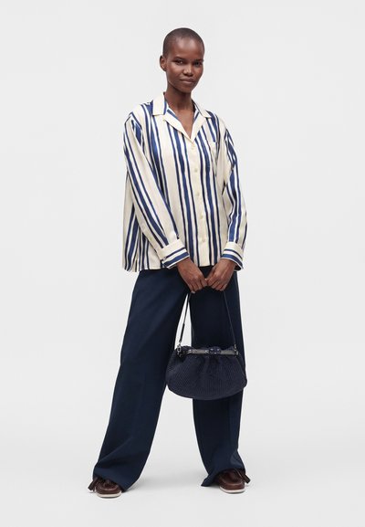 Personne portant une chemise ample crème avec des rayures verticales bleues, un pantalon large bleu marine, des chaussures marron et tenant un sac à main tricoté bleu marine.