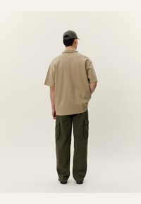 Beige kortermet skjorte med avslappet passform, sammen med mørkegrønn cargo-bukser med sidelommer, sett fra baksiden.