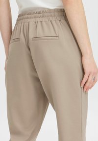 Beige, elastische Hosen mit glattem Textur, ausgestattet mit zwei Gesäßtaschen und einem lässigen Schnitt. Sichtbare Nähte entlang der Kanten.