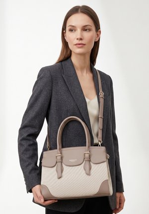 Vrouw in een donker colbert die een beige en taupekleurige gestructureerde handtas met bovenhandvatten en een verstelbare schouderriem vasthoudt.