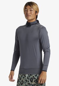 Grå långärmad träningsskjorta med hög krage och släta sömmar, tillverkad av slät, elastisk tyg. Bärs med mönstrade gröna shorts.