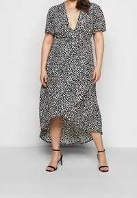 Czarna i biała sukienka maxi w grochy z głębokim dekoltem w kształcie litery V, krótkimi rękawami, talią podkreśloną paskiem i asymetrycznym dołem. Noszona z czarnymi sandałkami na obcasie z paskami.