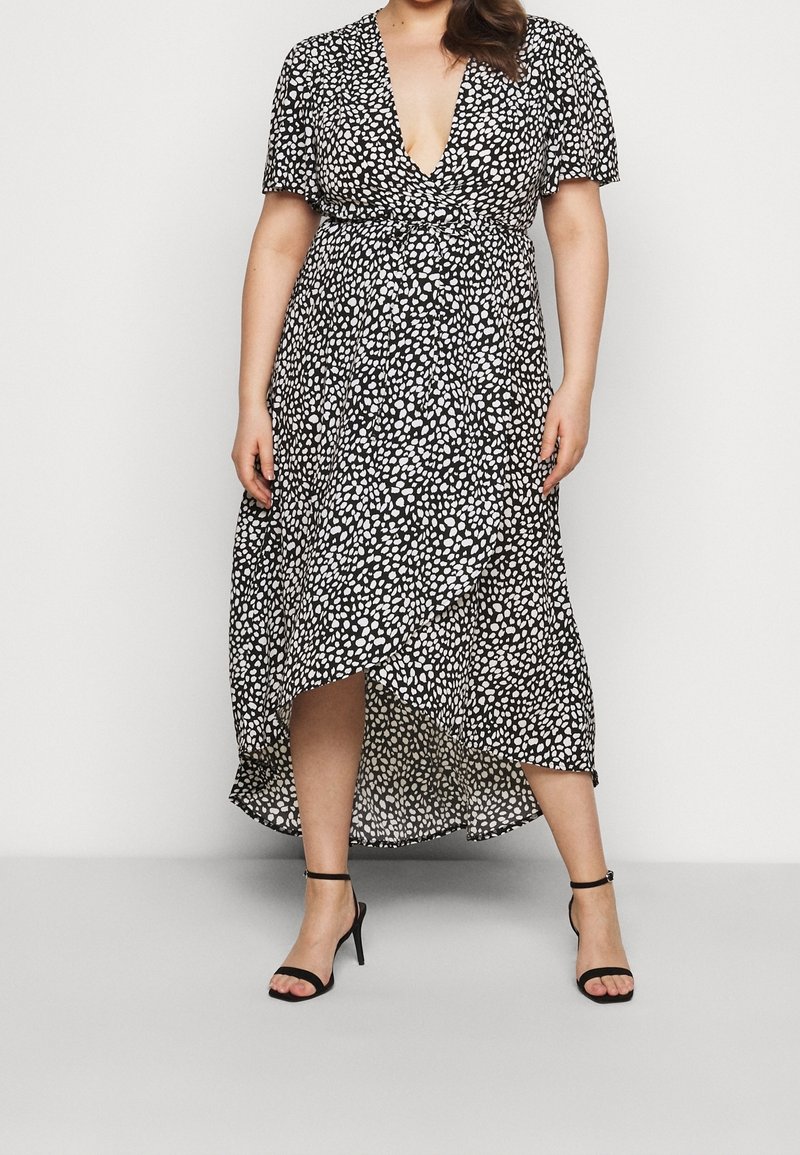 Czarna i biała sukienka maxi w grochy z głębokim dekoltem w kształcie litery V, krótkimi rękawami, talią podkreśloną paskiem i asymetrycznym dołem. Noszona z czarnymi sandałkami na obcasie z paskami.