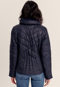 Marineblaue Steppjacke mit Zickzacksteppung, hohem Kragen und gerippten Handgelenkabschlüssen. Glatte, glänzende Textur mit tailliertem Schnitt.