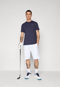 Tengerészkék rövid ujjú póló Calvin Klein logóval, fehér rövidnadrág, fehér golfcipő tengerészkék középpel, és golfütőt tart a kezében.