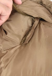 Interno di una giacca trapuntata marrone che mostra una tasca con zip realizzata in un tessuto liscio e lucido, con cuciture rinforzate.