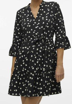 Robe noire à étages avec motifs de cœurs blancs, col en V, manches 3/4 volantées et ceinture nouée à la taille portée par une personne.