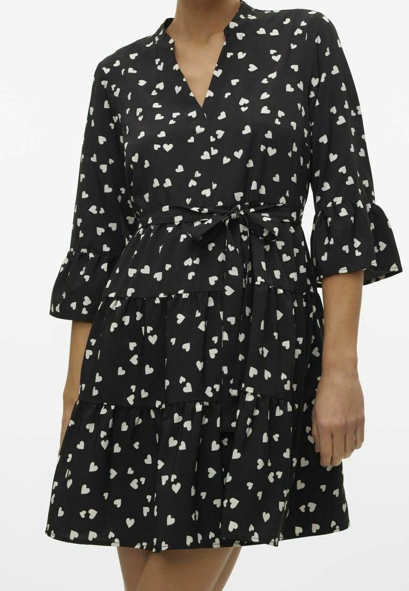 Robe noire à étages avec motifs de cœurs blancs, col en V, manches 3/4 volantées et ceinture nouée à la taille portée par une personne.