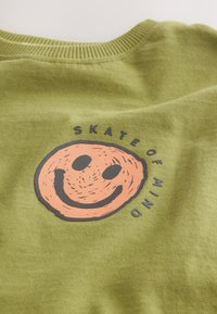 Olivově zelená látka s kulatým oranžovým smajlíkem a černým textem "SKATE OF MIND", uspořádaným kolem grafiky.