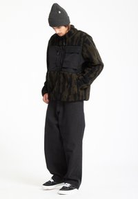Fleece-takki mustilla nailon-paneeleilla, jossa on khaki- ja musta tiikeriraidallinen kuvio, yhdistettynä leveisiin mustiin housuihin ja mustiin lenkkareihin.