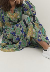 Abito lungo colorato con motivi floreali e geometrici in tonalità di verde e blu. Presenta maniche lunghe, scollatura a V e uno spacco laterale, abbinato a sneaker bianche.