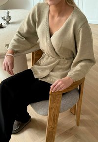 Beige gestrickte Wickeljacke mit V-Ausschnitt, weitem Ärmeln und einem Bindegurt, getragen über schwarzen Hosen. Modell sitzt auf einem Holzstuhl.