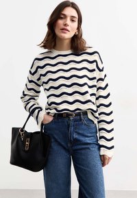 Marinblå och krämrandig stickad tröja med en vågmönster, kombinerad med mörkblå vida jeans och en strukturerad svart handväska med gulddetaljer.