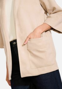 Ruka v kapse béžové bundy přes bílou halenku a tmavě modré džíny, ukazuje detail a texturu ležérního outfitu.