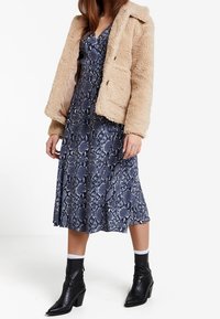 Beige Kunstfelljacke, knielanges Kleid in blauem Schlangenmuster, schwarze Stiefeletten mit leichtem Absatz und weiße Socken. Texturiert und geschichtet.