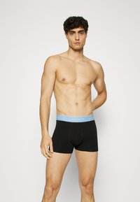 Schwarze Boxer-Shorts mit einem hellblauen elastischen Bund, auf dem das "Lacoste"-Logo zu sehen ist, aus einem glatten, dehnbaren Stoff gefertigt.
