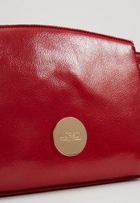 Borsa in ecopelle rossa con texture liscia, dotata di un logo circolare dorato e dettagli di cucitura lungo i bordi. Design compatto.
