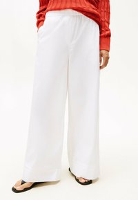 Pantaloni bianchi a gamba larga con vita elastica, due tasche laterali e orlo corto. Indossati con sandali neri a piedi nudi.
