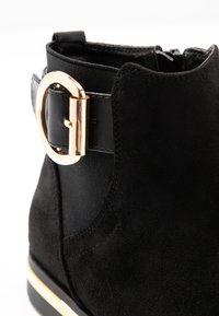 Botín negro hasta el tobillo con un cuerpo de ante texturizado, panel elástico y un acento de hebilla en tono dorado en una correa negra.