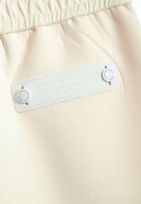 Pantalones con cintura elástica en suave tejido crema que presentan una etiqueta texturizada con letras en relieve y dos botones decorativos para el cierre.