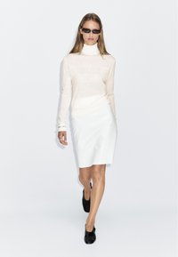 Pull à col roulé beige clair en tricot, jupe blanche ajustée et chaussures noires. Lunettes de soleil à monture étroite. Textures lisses et silhouette simple.