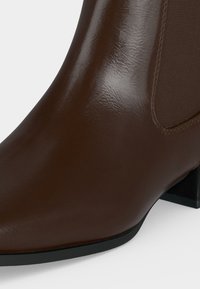 Bottine en cuir marron avec panneau latéral élastique et petit talon bloc noir sur fond blanc.