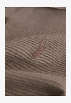 Barna szövött anyag texturált mintázattal, amelyen egy halvány rózsaszín árnyalatú szarvas logó van hímezve. Simított felület, melyen nincs jelentős mintázat.