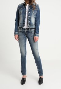 Jaschka dżinsowa z jasnym spranieniem, zapięcie na guziki z przodu, dwie kieszenie na piersi, połączona z dopasowanymi niebieskimi dżinsami i czarnymi butami wsuwanymi. Odzież codzienna.